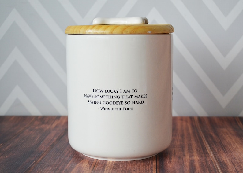 Puede incluir: Tarro de cer&aacute;mica blanco con tapa de madera y pomo en forma de hueso. El tarro tiene una cita de Winnie de Pooh que dice: "Qu&eacute; suerte tengo de tener algo que hace que decir adi&oacute;s sea tan dif&iacute;cil. - Winnie de Pooh"
