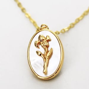Puede incluir: Un collar de cadena dorada con un colgante ovalado blanco con un diseño de flor dorada.
