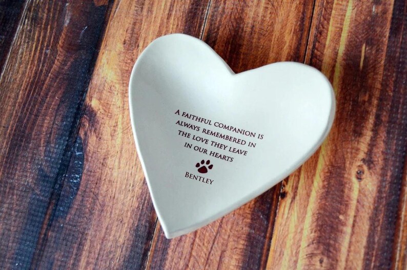 May include: White ceramic heart-shaped dish with the inscription "A faithful companion is always remembered in the love they leave in our hearts" and the name "Bentley" below a paw print.