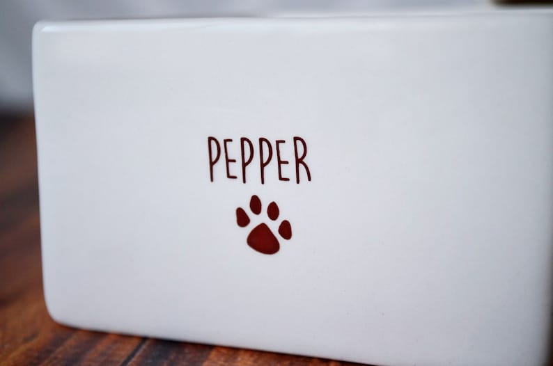 K&ouml;nnte beinhalten: Wei&szlig;e Keramik-Futternapf f&uuml;r Haustiere mit dem Namen "Pepper" und einem roten Pfotenabdruck.