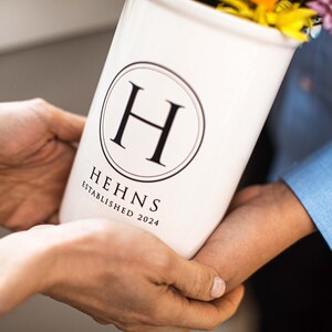 Puede incluir: Un jarr&oacute;n de cer&aacute;mica blanco con un logotipo circular negro con la letra "H" y el texto "HEHNS ESTABLISHED 2024". El jarr&oacute;n es sostenido por dos personas y est&aacute; lleno de flores coloridas.