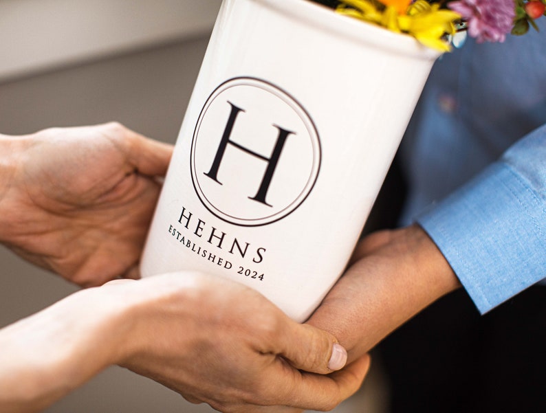 Puede incluir: Un jarr&oacute;n de cer&aacute;mica blanco con un logotipo circular negro con la letra "H" y el texto "HEHNS ESTABLISHED 2024". El jarr&oacute;n es sostenido por dos personas y est&aacute; lleno de flores coloridas.