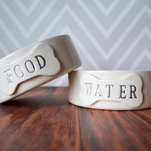 May include: Two off-white ceramic pet bowls with a glossy finish. One bowl is tilted and features a raised bone-shaped plaque with the word FOOD embossed in black. The second bowl sits flat and features a bone-shaped plaque with WATER embossed in black.