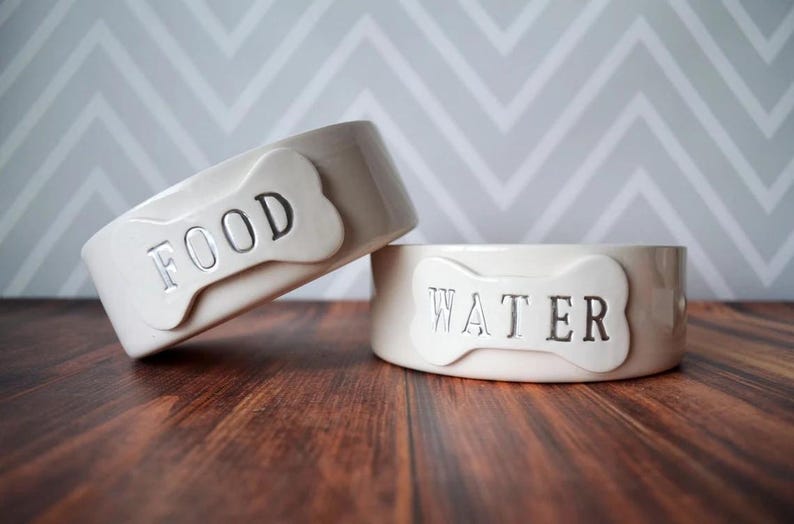 May include: Two off-white ceramic pet bowls with a glossy finish. One bowl is tilted and features a raised bone-shaped plaque with the word FOOD embossed in black. The second bowl sits flat and features a bone-shaped plaque with WATER embossed in black.