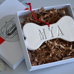 An off-white, ceramic, dog bone-shaped ornament with the pet name "MYA" stamped into the surface. The name is painted in metallic silver. The ornament has a red cord and is shown in a white gift box on silver background.