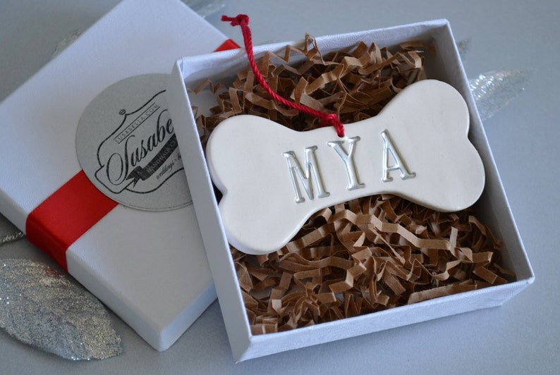 An off-white, ceramic, dog bone-shaped ornament with the pet name "MYA" stamped into the surface. The name is painted in metallic silver. The ornament has a red cord and is shown in a white gift box on silver background.