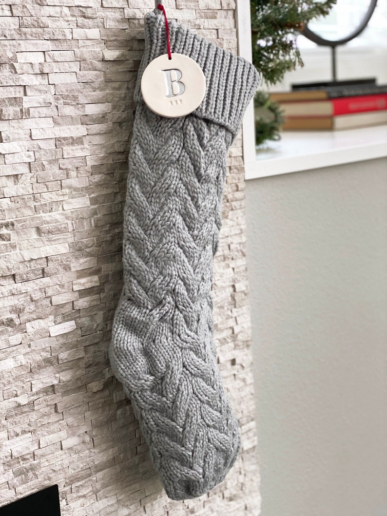 A grey knitted holiday stocking with off-white, round, ceramic ornament hanging from red cord. The ornament is stamped with an initial and name. The stocking is hung against a white brick fireplace.