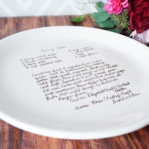 A large round serving plate sits on a wooden table with flowers. The plate has light brown/sepia text printed on the front in a handwritten script font. The plate is shown from a side angle, showing a slight rim and flat shape.