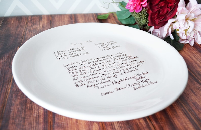 A large round serving plate sits on a wooden table with flowers. The plate has light brown/sepia text printed on the front in a handwritten script font. The plate is shown from a side angle, showing a slight rim and flat shape.