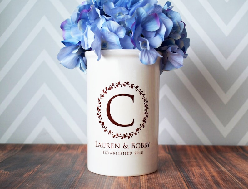 May include: White ceramic vase with a brown wreath and the letter C in the center. The vase has the text "Lauren & Bobby Established 2018" printed on the bottom. A bouquet of blue hydrangeas is in the vase.