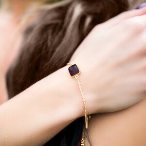A delicate gold chain bracelet with a square-shaped dark ruby gemstone pendant.