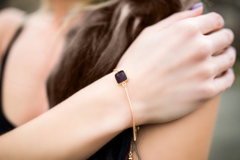 A delicate gold chain bracelet with a square-shaped dark ruby gemstone pendant.