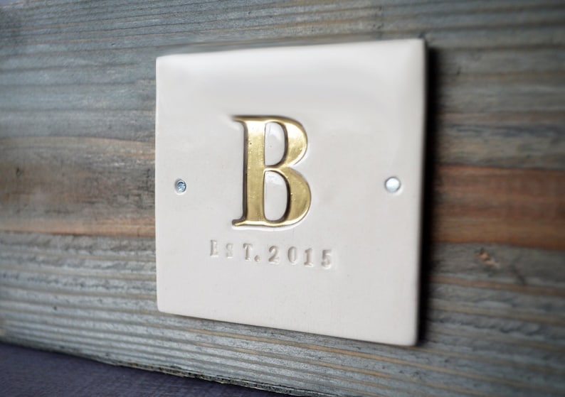 A white ceramic tile on a wood planter box (wedding centerpiece) with hand-stamped initial (painted gold) and Est. date.