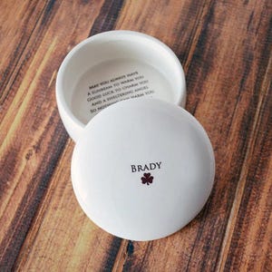 May include: A white ceramic trinket box with a lid, resting on a wooden surface. The lid is inscribed with the name "Brady" and a small shamrock. The inside of the box has an inscription. The box is open, revealing the interior.