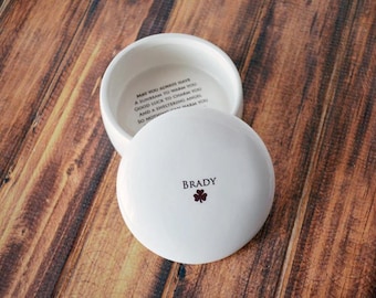May include: A white ceramic trinket box with a lid, resting on a wooden surface. The lid is inscribed with the name "Brady" and a small shamrock. The inside of the box has an inscription. The box is open, revealing the interior.