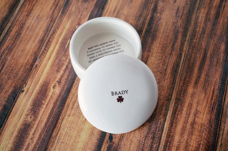 May include: A white ceramic trinket box with a lid, resting on a wooden surface. The lid is inscribed with the name "Brady" and a small shamrock. The inside of the box has an inscription. The box is open, revealing the interior.