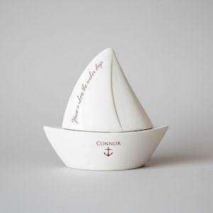 Op de afbeelding: Een witte keramische zeilbootvormige container met een zeil en bootbasis. Het zeil heeft de tekst "Home is where the anchor drops" in donkerrood schrift. De bootbasis heeft de naam "Connor" en een rode anker.