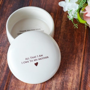 May include: White ceramic trinket box with a lid. The lid has the inscription "All that I am I owe to my mother" with a small heart symbol. The inside of the box has the inscription "Thank you for your unconditional love".