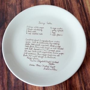 A large round serving plate sits on a wooden table, shown from the top. The plate has centered light brown/sepia text printed on the front in a handwritten script font. The text is a custom handwritten family recipe. The plate has a slight rim.