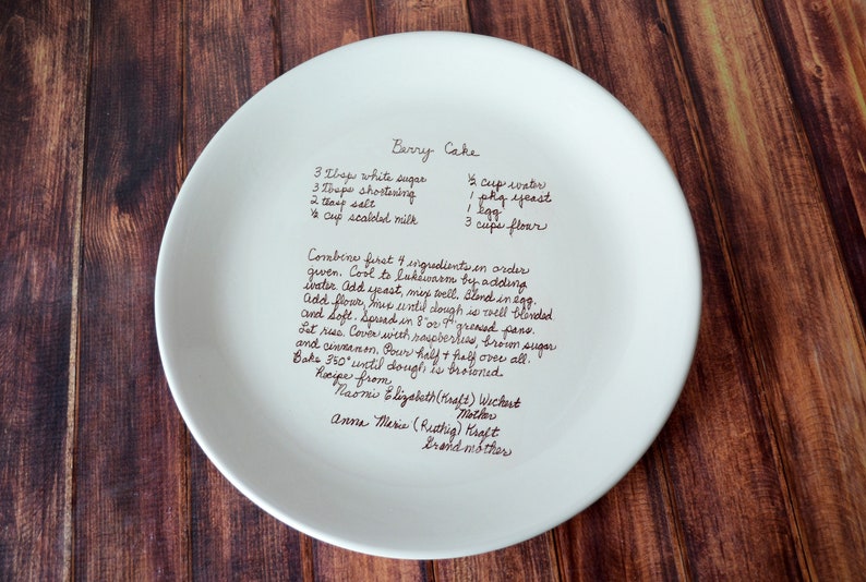 A large round serving plate sits on a wooden table, shown from the top. The plate has centered light brown/sepia text printed on the front in a handwritten script font. The text is a custom handwritten family recipe. The plate has a slight rim.