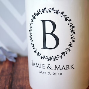 May include: White ceramic jar with a black wreath design and the letter "B" in the center. The text "Jamie & Mark" and "May 5, 2018" is printed below the wreath.