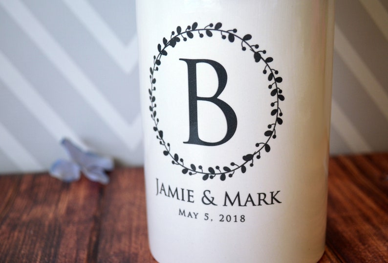 May include: White ceramic jar with a black wreath design and the letter "B" in the center. The text "Jamie & Mark" and "May 5, 2018" is printed below the wreath.