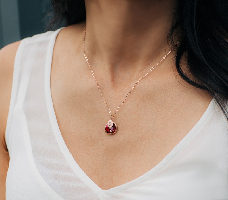 May include: A rose gold necklace with a teardrop-shaped pendant featuring a red gemstone and the letter "S" engraved on the front.