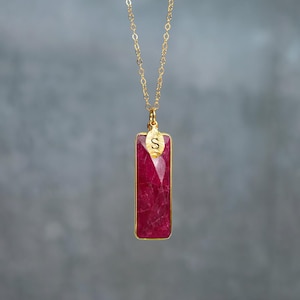 May include: A gold chain necklace featuring a rectangular, faceted, deep pink gemstone pendant. A small, gold, leaf-shaped charm is attached to the top of the pendant, engraved with the capital letter S. The pendant is framed in a thin gold border.