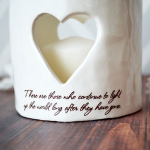 Close up image of an off white ceramic votive with two lines of centered light brown sepia script text below a heart on the front. Text reads There are those who continue to light up the world long after they have gone.