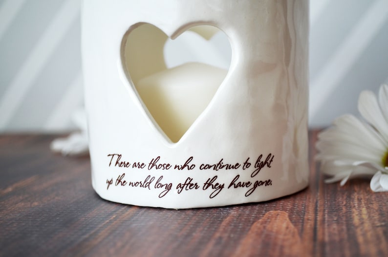 Close up image of an off white ceramic votive with two lines of centered light brown sepia script text below a heart on the front. Text reads There are those who continue to light up the world long after they have gone.