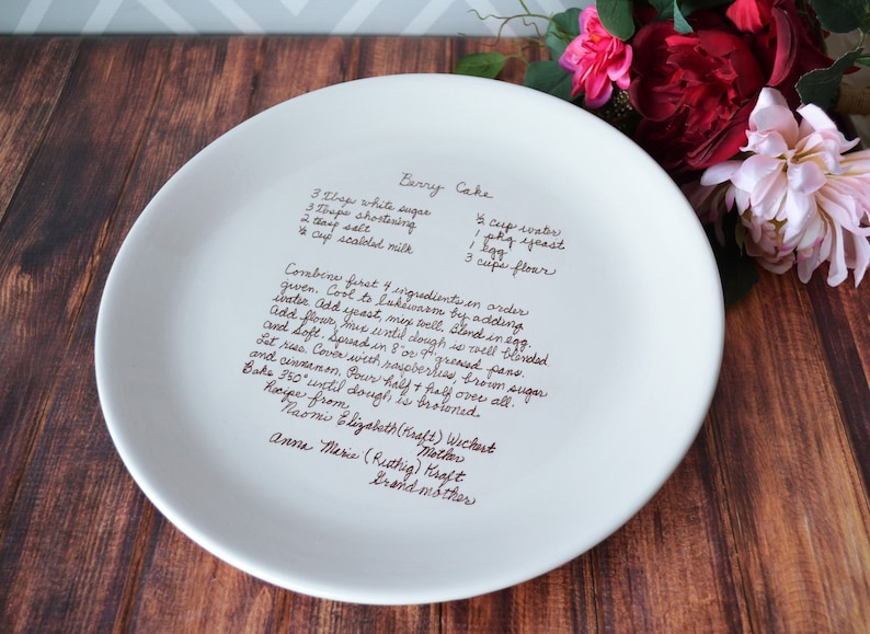 A large round serving plate sits on a wooden table with flowers. The plate has light brown/sepia text printed on the front in a handwritten script font. The text is a custom handwritten family recipe.
