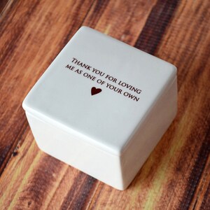 May include: A square, off-white keepsake box with a lid. The lid features the text "THANK YOU FOR LOVING ME AS ONE OF YOUR OWN" in dark red, with a small heart symbol. The box is set against a wooden background.