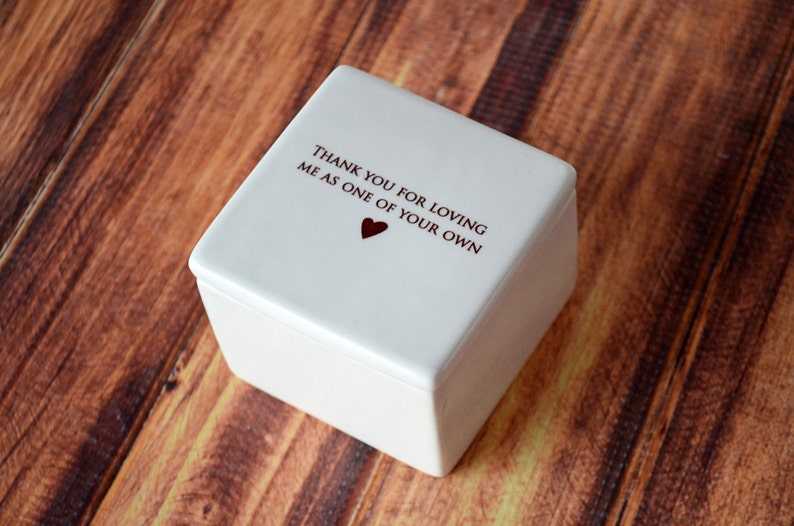 May include: A square, off-white keepsake box with a lid. The lid features the text "THANK YOU FOR LOVING ME AS ONE OF YOUR OWN" in dark red, with a small heart symbol. The box is set against a wooden background.