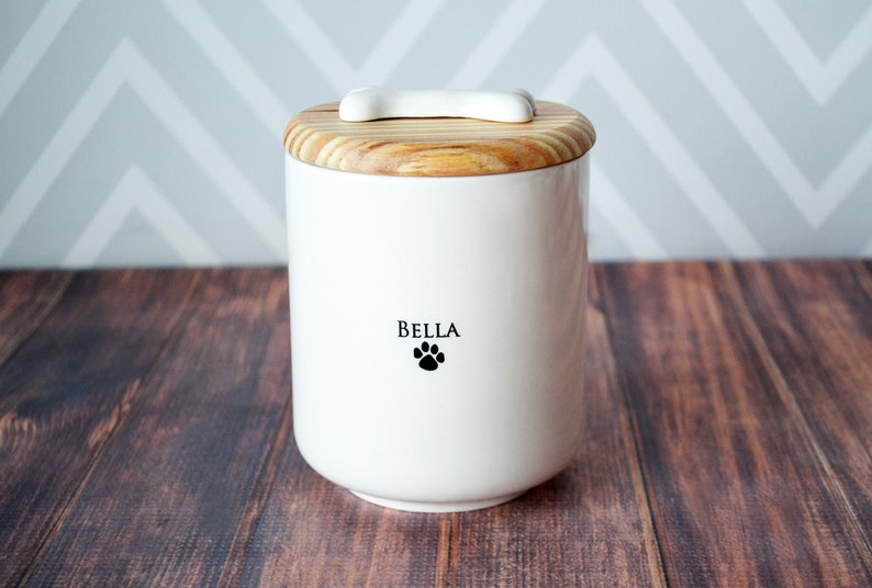 White ceramic pet food container with a wooden lid and a bone-shaped handle. The container has a black paw print and the name 'BELLA' printed on the front.