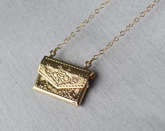 May include: A gold necklace with a detailed, ornate, envelope-shaped pendant. The pendant has a floral design and is attached to a delicate chain.