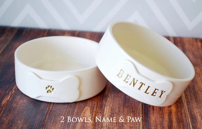 Image of the 2 bowls, name & paw option. Two bowls are shown, each with a bone shaped tile on the front. One bowl tile is stamped with a pet name in all capital letters, the other has a paw print image, both painted in metallic gold.
