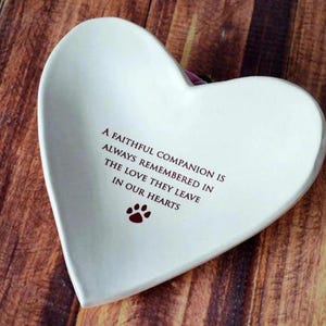 May include: White ceramic heart-shaped dish with a red paw print and the inscription "A faithful companion is always remembered in the love they leave in our hearts."