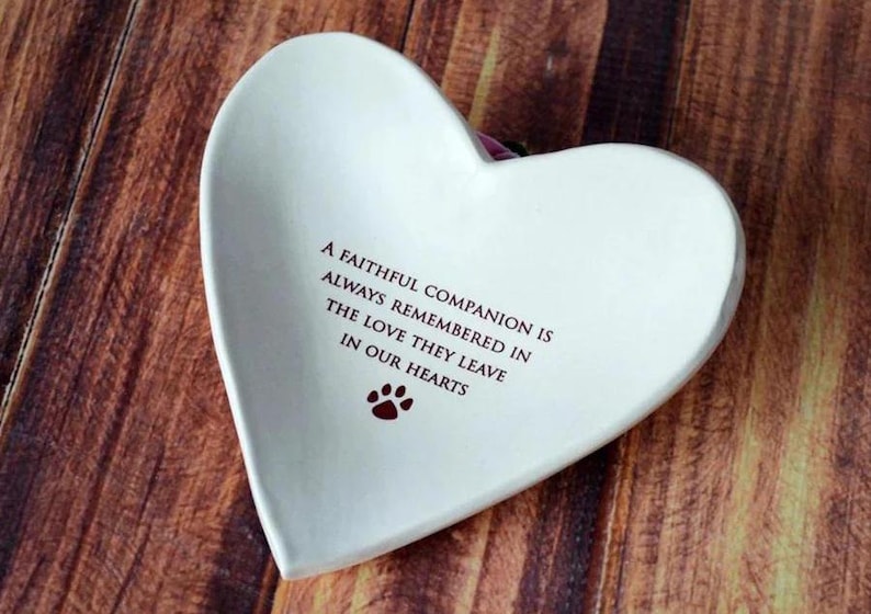May include: White ceramic heart-shaped dish with a red paw print and the inscription "A faithful companion is always remembered in the love they leave in our hearts."