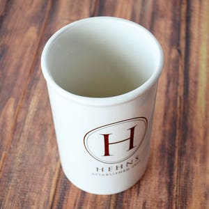 Puede incluir: Un recipiente blanco de cer&aacute;mica para utensilios de cocina, de forma cil&iacute;ndrica. Tiene un dise&ntilde;o ovalado marr&oacute;n con la letra "H" y el texto "HEHNS ESTABLISHED 2013". El recipiente est&aacute; sobre un fondo de madera.