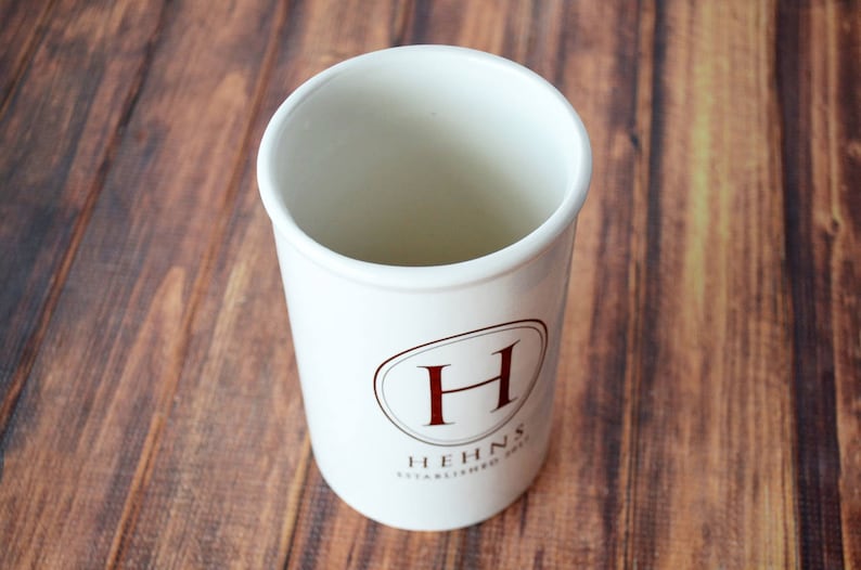 Puede incluir: Un recipiente blanco de cer&aacute;mica para utensilios de cocina, de forma cil&iacute;ndrica. Tiene un dise&ntilde;o ovalado marr&oacute;n con la letra "H" y el texto "HEHNS ESTABLISHED 2013". El recipiente est&aacute; sobre un fondo de madera.