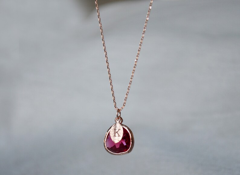 A pinkish red ruby colored transparent glass stone charm is framed in rose gold metal and hangs from a rose gold link chain. There is a small leaf shaped charm with the letter K stamped on the front, hanging in front of the ruby charm.