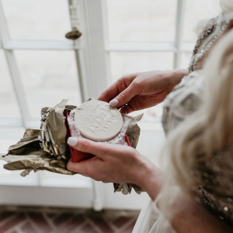 Image of an off-white gloss-glazed ceramic round-shaped ornament. The ornament is being unwrapped from a red gift box at a wedding by the bride.