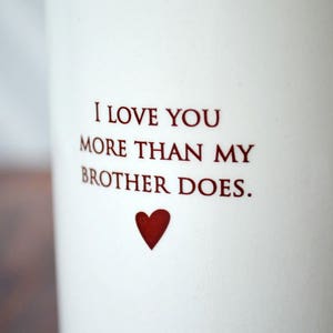 White ceramic mug with the text "I love you more than my brother does."  and a heart.