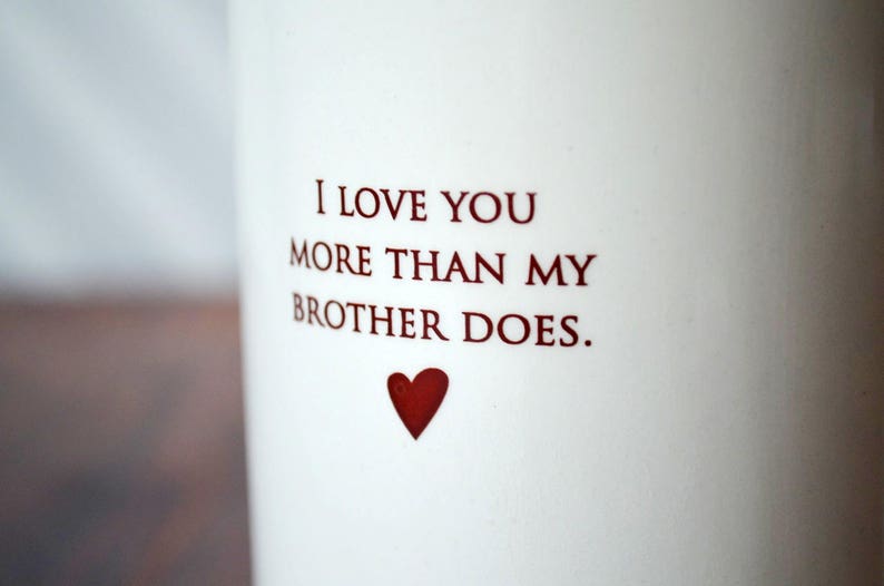 White ceramic mug with the text "I love you more than my brother does."  and a heart.