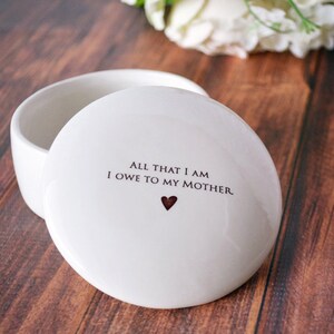 May include: White ceramic trinket box with a lid. The lid has the text "All that I am I owe to my mother." with a small red heart.