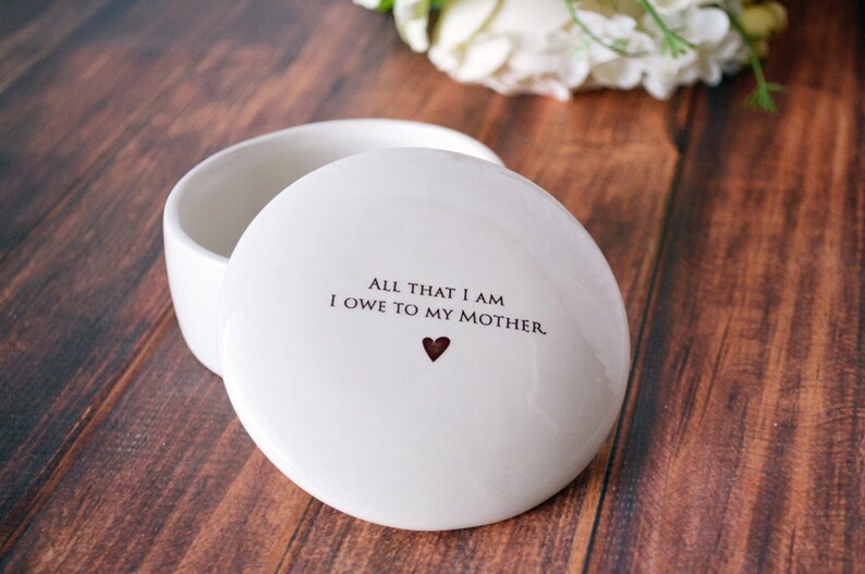 May include: White ceramic trinket box with a lid. The lid has the text "All that I am I owe to my mother." with a small red heart.