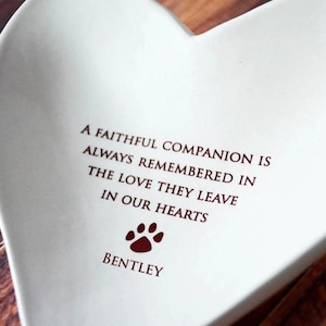 May include: White ceramic heart-shaped dish with a red paw print and the inscription "A faithful companion is always remembered in the love they leave in our hearts" and the name "Bentley" below the paw print.