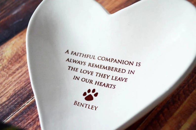 May include: White ceramic heart-shaped dish with a red paw print and the inscription "A faithful companion is always remembered in the love they leave in our hearts" and the name "Bentley" below the paw print.