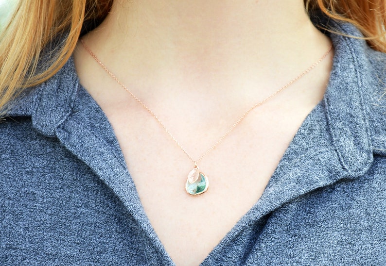 The necklace is shown on a model wearing a grey button down shirt. A rose gold metal chain with aquamarine charm and leaf charm hangs around the model's neck. The aquamarine is framed in rose gold and the leaf on top is stamped with an initial.