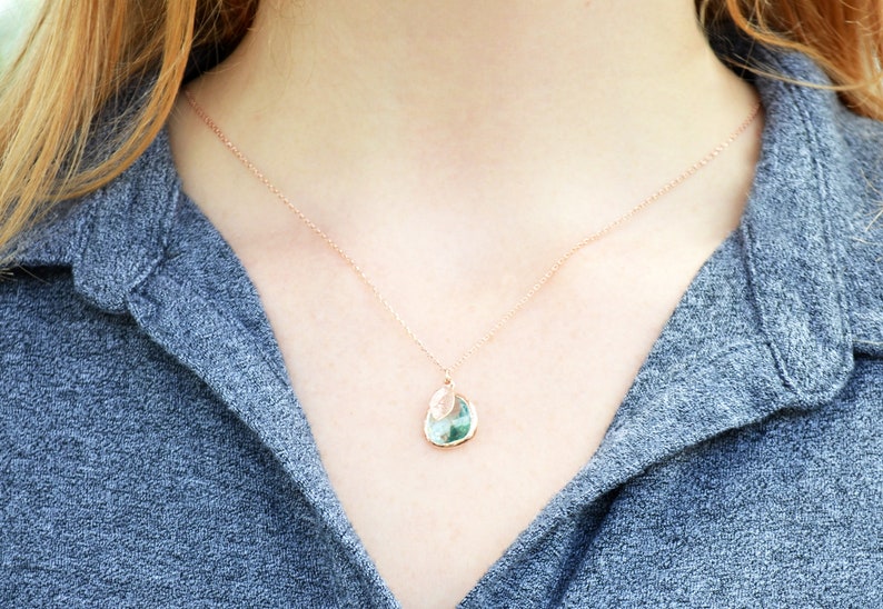 The necklace is shown on a model wearing a grey button down shirt. A rose gold metal chain with aquamarine charm and leaf charm hangs around the model's neck. The aquamarine is framed in rose gold and the leaf on top is stamped with an initial.